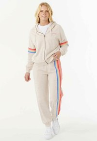 Vrouw in een beige hoodie met rits en sweatpants met regenboogstrepen langs de mouwen en broekspijpen, staand en naar voren lopend.