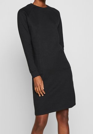 Kvinde iført en sort, afslappet kjole med lange ærmer og knælængde, med to forlommer, mod en ensartet lys baggrund.