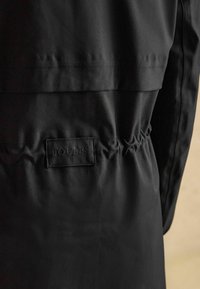 Čierna bunda s hladkým, matným povrchom. Obsahuje elastické detaily v oblasti pása a tkaný štítok "JOULES" prišitý na zadnej strane.