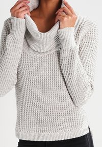 Jasnoszary dzianinowy sweter z dekoltem w stylu cowl, teksturowanym prążkowaniem i długimi rękawami. Posiada dopasowany fason w talii i lekko luźne mankiety.
