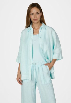 Frau trägt ein lockeres hellblaues Hemd mit Knöpfen, passende Hosen mit Kordelzug und ein Tanktop, steht mit einer Hand in der Tasche.