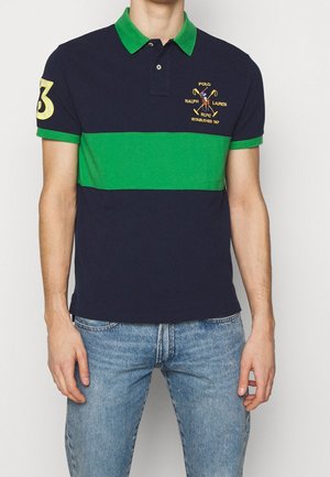 Mann trägt ein dunkelblau-grün gestreiftes Poloshirt mit gesticktem Logo und der Nummer 3 auf dem Ärmel, kombiniert mit blauen Jeans.