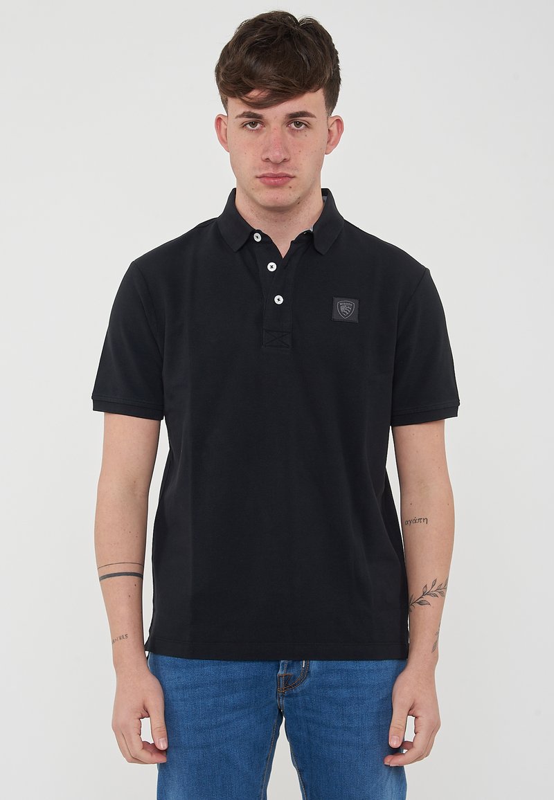 Polo nero in cotone, dotato di colletto con tre bottoni, maniche corte e una piccola toppa con logo sul petto.