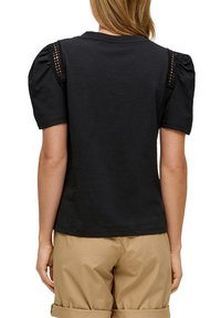 Zwarte t-shirt met puff mouwen en kantdetails op de schouders, gemaakt van zachte stof. Gecombineerd met beige broek, gezien van achteren.