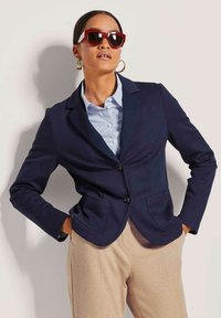 Marineblazer mit zwei Vordertaschen, Ein-Knopf-Verschluss und Revers; getragen über einem hellblauen gestreiften Hemd und beigen Hosen.