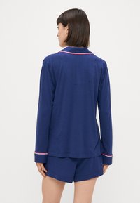 Tamsiai mėlynos spalvos pižamų rinkinys su ilgomis rankovėmis marškiniais ir šortais, su kontrastingais raudonais siūlėmis ant manžetų ir kaklo, pagamintas iš lengvos medžiagos.