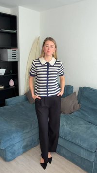 Γυναίκα που στέκεται με τα χέρια στις τσέπες, φοράει ριγέ κοντομάνικη μπλούζα και μαύρο παντελόνι σε σαλόνι με μπλε καναπέ.