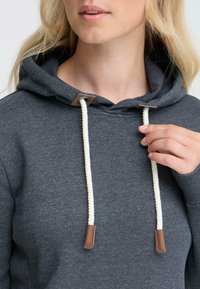 Un sweat à capuche gris avec une texture douce, doté d'un cordon de serrage en crème et de pointes en cuir marron. La capuche est doublée d'un tissu assorti.