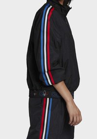 Veste de sport noire avec des rayures rouges, bleues et blanches sur les manches et une ceinture élastique ornée de détails de logo. Matière coupe-vent, fermeture éclair.