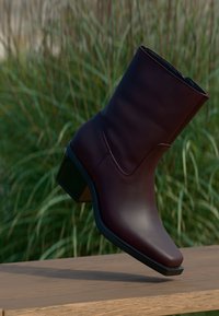 Dark brown mid-calf leather boot with block heel balancing on a wooden surface, green tall grass blurred in the background.