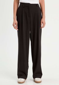 Pantaloni neri a gamba larga realizzati in un tessuto liscio con vita alta, dotati di pieghe frontali e tasche laterali, abbinati a sneaker bianche.