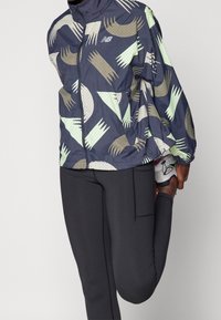 Czarna i szara kurta przeciwwiatrowa z abstrakcyjnymi kształtami i liniami, zapinana na zamek, pod spodem czarne sportowe legginsy.