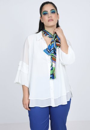 White, button-up blouse with sheer, layered sleeves; paired with blue pants and a multicolored, striped scarf tied at the neck.