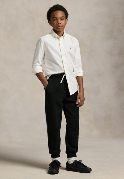 Garçon debout, une main dans la poche, portant une chemise blanche boutonnée, un pantalon noir à cordon, des chaussettes blanches et des baskets noires sur fond uni.