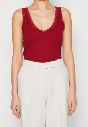 Vrouw draagt een mouwloze, dieprode top met een V-hals en kanten rand, ingestopt in wijde, witte broek met hoge taille en lussen voor een riem.