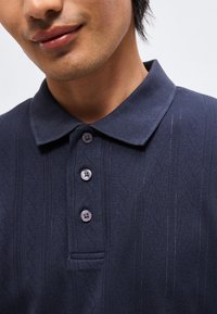 Άνδρας που φοράει μπλε σκούρο πλεκτό πόλο με γιακά και τρία σκουρόχρωμα κουμπιά, εμφανιζόμενος από τον λαιμό μέχρι το μέσο του στήθους με λευκό φόντο.