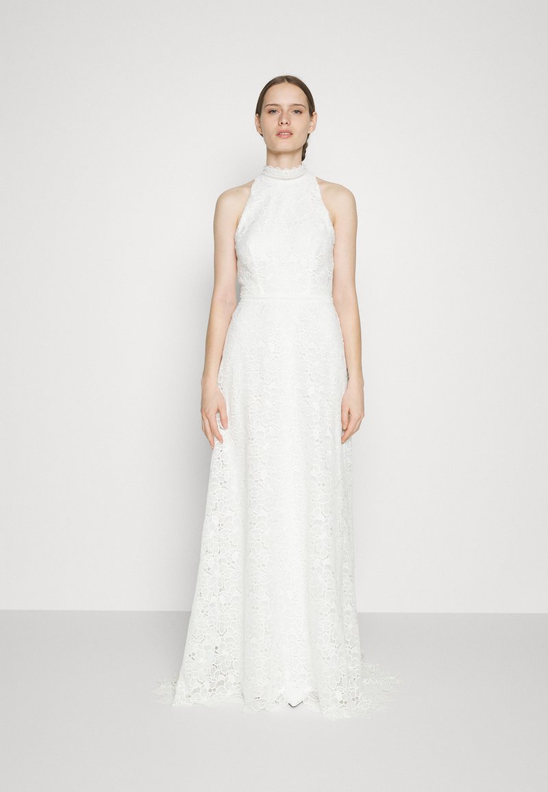 Mujer vestida con un vestido sin mangas de encaje blanco de cuello alto con patrones florales, de pie contra un fondo gris claro liso.