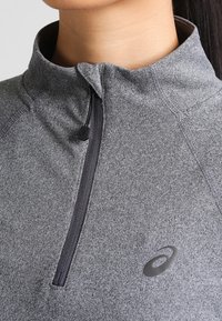 Grauer Reißverschlusspullover aus weichem, strukturiertem Stoff, mit Stehkragen und schwarzem Reißverschluss, sowie einem kleinen Logo im Brustbereich.