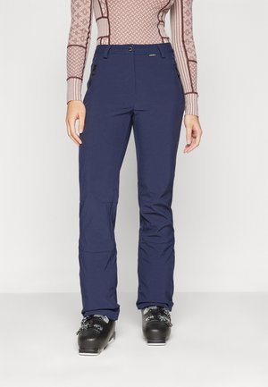 Vrouw die marineblauwe skibroek, zwarte skischoenen en een patroonrijke lange mouw top draagt, staand tegen een effen lichte achtergrond.