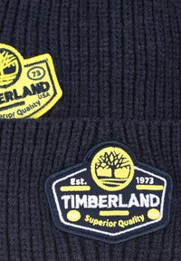 Twee donkerblauwe gebreide mutsen met gele en witte Timberland-logo patches, waarop een boom staat en de tekst "Superieure Kwaliteit Est. 1973 VS."