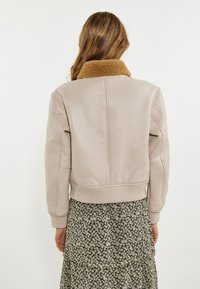 Beige læderjakke med en brun shearling-krave, ribbede manchetter og kant. Båret over en grøn blomstermønstret kjole, der viser et lagdelt design.