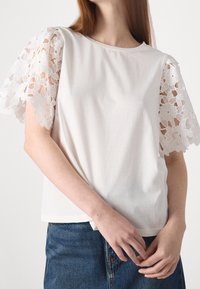 Biały top z krótkim rękawem z misternymi koronkowymi rękawami w kwiatowy wzór. Główna część wykonana z gładkiej bawełny, o luźnym kroju i okrągłym dekolcie. Widoczny deninowy dół.