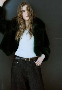 Zielona kurtka z imitacji futra na białym prążkowanym topie, zestawiona z szerokimi spodniami z ciemnego denimu i brązowym paskiem; długie falujące włosy częściowo zasłaniają twarz.