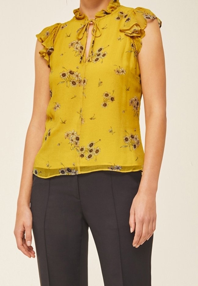 Blouse jaune sans manches avec petit imprimé floral, épaules à volants, col avec lien, associée à un pantalon noir cintré.