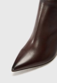 Botín de tobillo de cuero marrón con punta afilada, textura suave y silueta estilizada. Presenta un diseño elegante y un tacón de aguja.