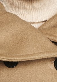 Beige uldfrakke med rund krave, dobbelte knapper og blød tekstur. En cremefarvet ribbet sweater er delvist synlig under.