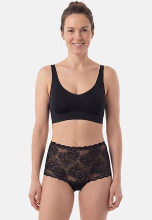 Donna con i capelli raccolti che indossa un reggiseno nero senza cuciture e mutandine nere in pizzo a vita alta, sorridendo su uno sfondo bianco.