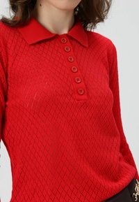 Rode gebreide trui met een gestructureerd ruitpatroon, voorzien van een kraag en zes bijpassende knopen aan de voorkant.