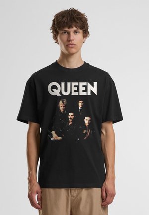 Jeune personne aux cheveux courts et bouclés portant un T-shirt noir du groupe Queen avec une photo des quatre membres du groupe.