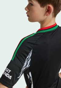 Svart sportströja med röda och gröna ränder på kragen och axlarna, texturerade detaljer och ett vitt grafiskt inslag.
