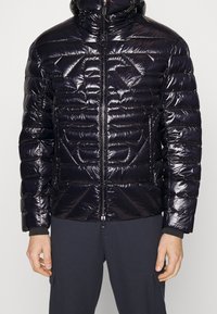 Lesklá černá prošívaná bunda s vysokým límcem, předním zipem a geometrickými švy. Obsahuje elastické manžety a přiléhavý střih.