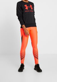 Felpa nera con logo rosso "Under Armour" e tasca, abbinata a leggings arancioni brillanti con accenti neri e motivo a pois. Sneakers nere.