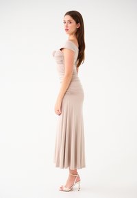 Lungo abito beige con corpetto aderente, design off-shoulder e gonna fluente, realizzato in tessuto morbido. Indossato con sandali con tacco alto.