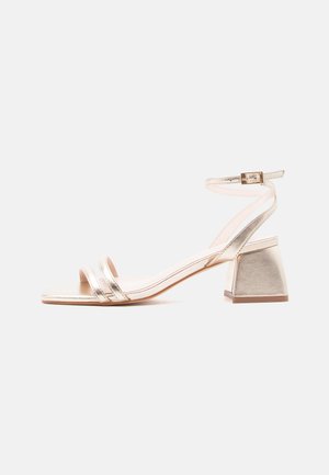 Sandale ouverte dorée pour femme avec petit talon bloc, double brides à l'avant et bride de cheville avec boucle, sur fond blanc.