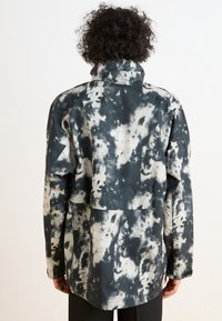 Czarna i biała kurtka tie-dye z wysokim kołnierzem, luźnym krojem i dużą tylną kieszenią. Posiada fakturowany materiał oraz subtelne logo marki.