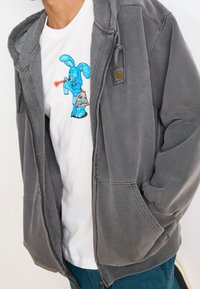 Szara bluza z rozpinanym zamkiem o miękkiej teksturze. Biała koszulka pod spodem ma graficzny nadruk niebieskiego kreskówkowego królika z puszką spreju.