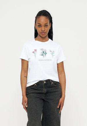 Γυναίκα με πλεξούδες φοράει λευκό T-shirt με ροζ, κίτρινα και λευκά λουλούδια και το κείμενο "STAY WILD, FLOWERCHILD", συνδυασμένο με μαύρο τζιν.