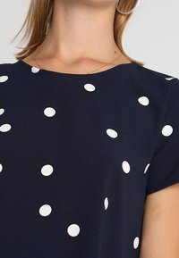 Marineblauw shirt met een ronde halslijn en korte mouwen, versierd met witte polka dots in verschillende maten. Zachte, gestructureerde stof.