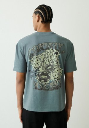 Mand med flettet hår iført en blågrøn t-shirt med et motiv af et skelet-hånd, terninger, bjørnefælder og teksten "Grateful Dead" på ryggen.