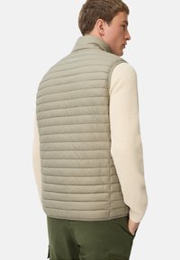Mann in beigefarbenem gesteppter ärmelloser Weste über einem cremefarbenen Strickpullover mit grünen Cargo-Hosen, mit dem Rücken zur Kamera und einer Hand in der Tasche.
