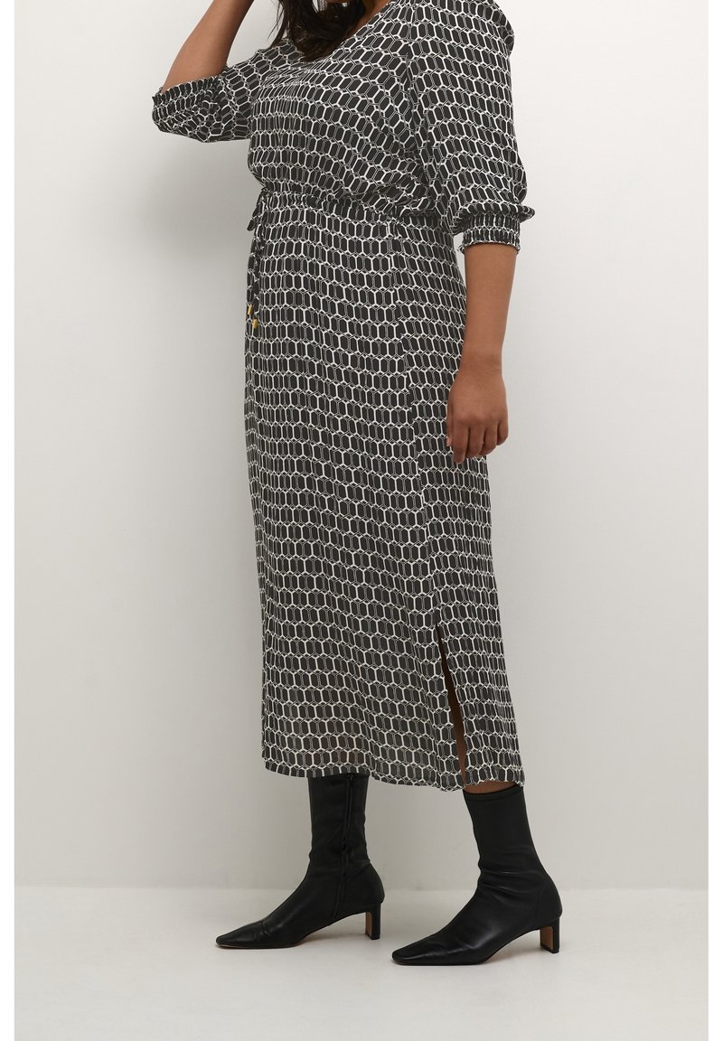 Femme portant une robe midi à motifs géométriques noir et blanc avec manches ballon, fente latérale, et bottines noires à talons moyens, debout contre un mur blanc.