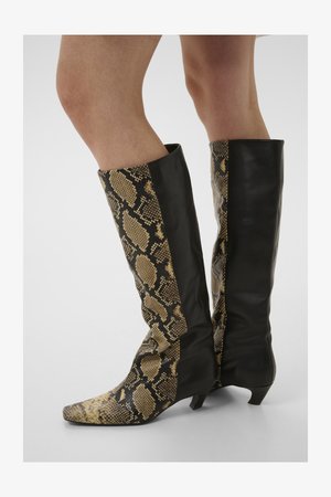 Põlveni ulatuvad saapad, millel on esiküljel beežide ja mustade madu naha mustriga ning taga must nahk, kantud paljaste jalgadega.