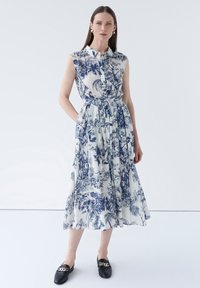 Robe à motif floral bleu et blanc, sans manches, avec des boutons sur le devant, taille cintrée, jupe longue, associée à des mocassins noirs.