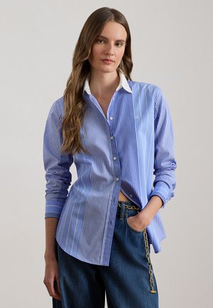 Pikkade pruunide juustega naine, kandes sinise ja valge triibulise nööbitava särgi valge krae ja tumesiniste teksadega, millel on kuldne kettvöö.