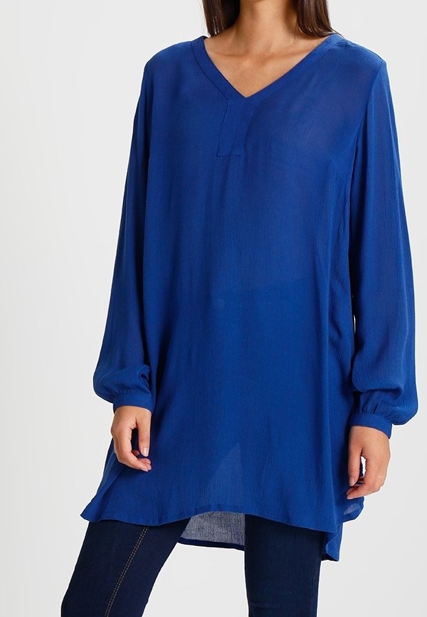 Femme portant une tunique ample à manches longues bleu roi avec un col en V, associée à un pantalon sombre ajusté, sur un fond uni.