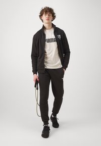 Tuta sportivo nero con giacca zip e pantaloni coordinati. Presenta un logo bianco sulla giacca e sui pantaloni, abbinati a una maglietta beige chiaro.